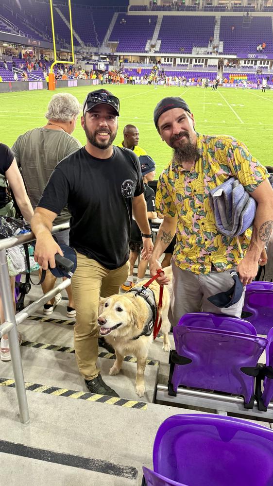 Two handlers standing with a service dog in training in a stadium concourse.